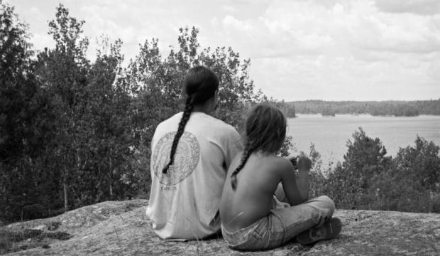 Black and white image. A man with a braid is sitting on a rock structure. He sits close to a boy with a braid. Their backs face us as they overlook a cliff and trees. 